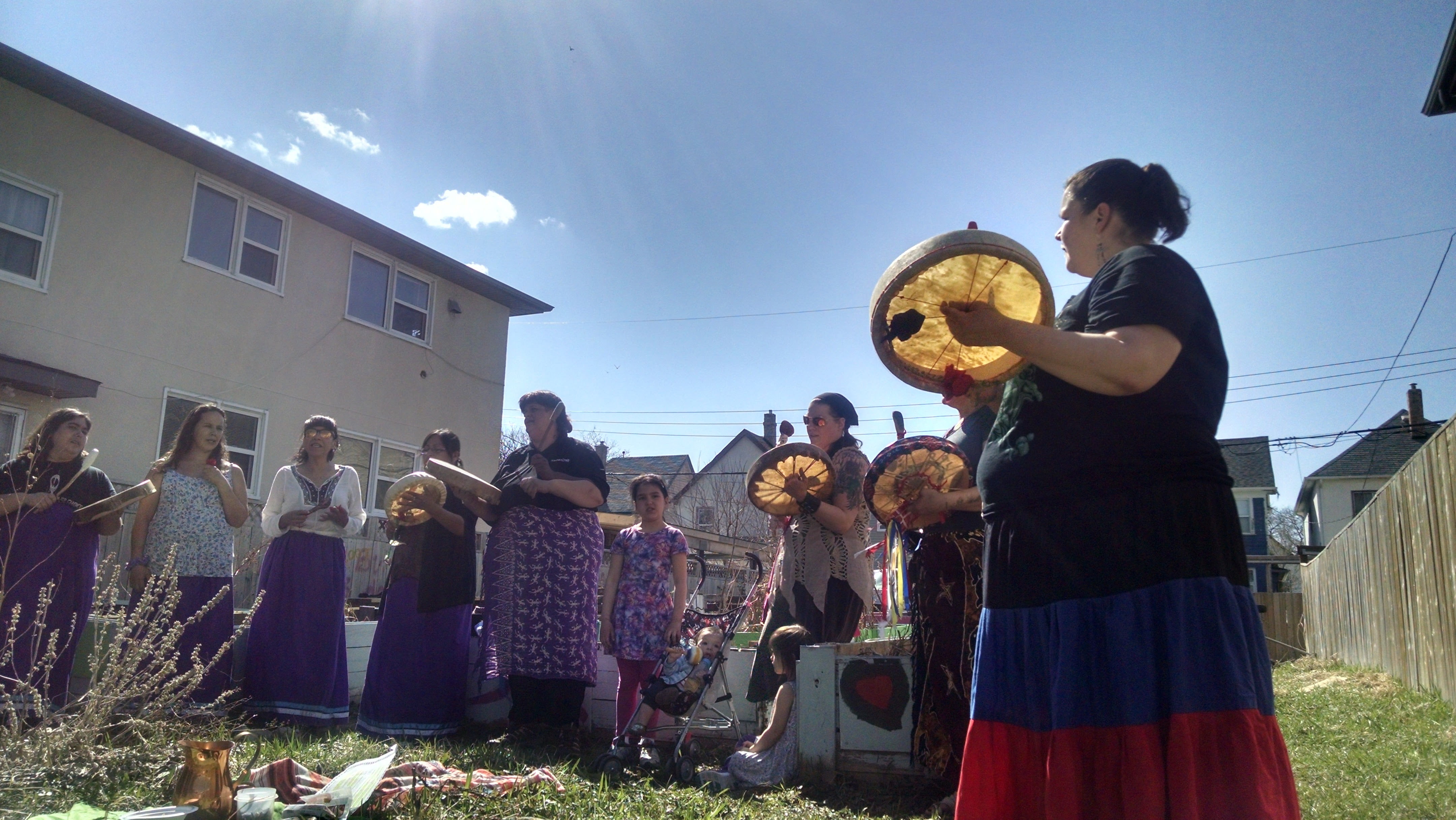 Group of women singing with hand drums in a community garden on a sunny day.